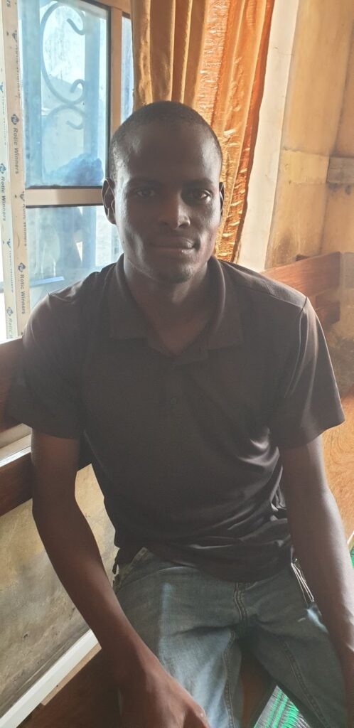 Man in a dark polo shirt sitting indoors with a window and curtains in the background.