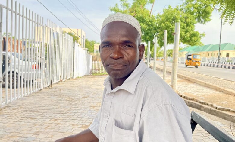 Man in a white hat smiling while sitting on a bench outdoors with trees and a vehicle in the background.