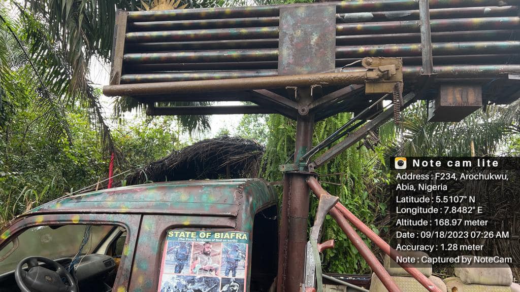 An old rusty vehicle with a poster and a nest on top under a metal structure in a tropical setting.