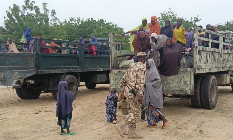 People in colorful clothing riding in open-back trucks with a soldier in the foreground.