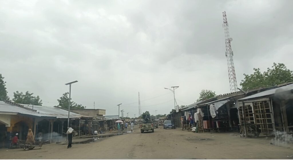 Dusty street with a few people, shops, vehicles, and a tall communication tower under an overcast sky.