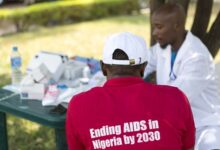 Man in a red shirt with "Ending AIDS in Nigeria by 2030" text, facing a healthcare worker at a table with medical supplies.