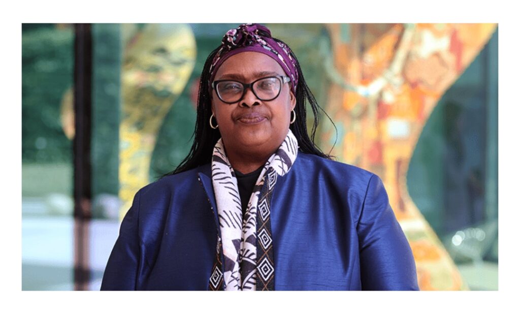 Woman with headscarf and glasses standing in front of colorful glass artwork.