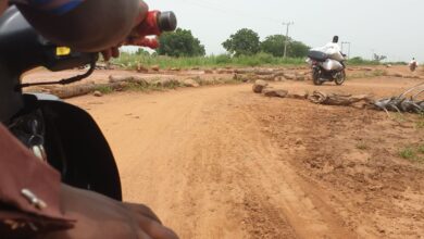 First-person view from a motorcycle approaching a dirt road intersection with a passing rider and scattered rocks.