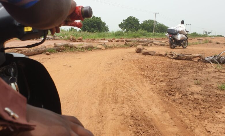 First-person view from a motorcycle approaching a dirt road intersection with a passing rider and scattered rocks.