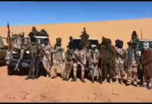 A group of people in desert attire posing in front of vehicles on a sandy terrain.