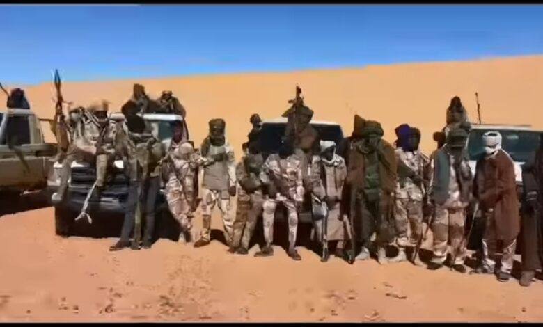 A group of people in desert attire posing in front of vehicles on a sandy terrain.