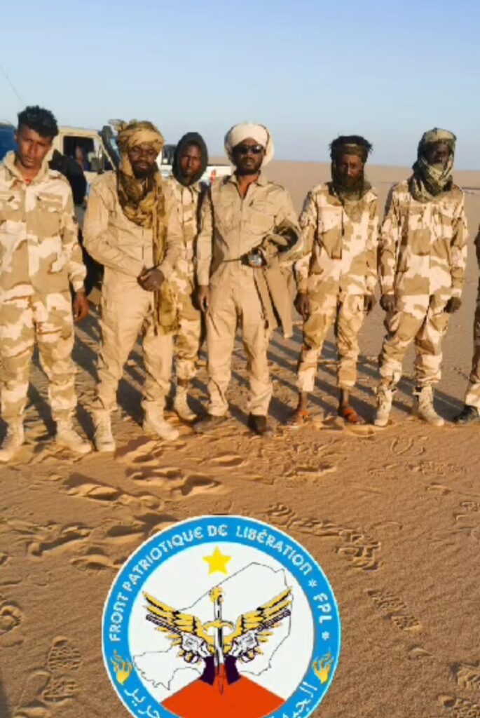 Group of six people in desert camo standing by a vehicle above a logo on the sand.