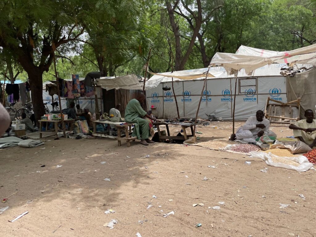 People sitting near makeshift tents with UNHCR logos in a tree-shaded, outdoor setting.