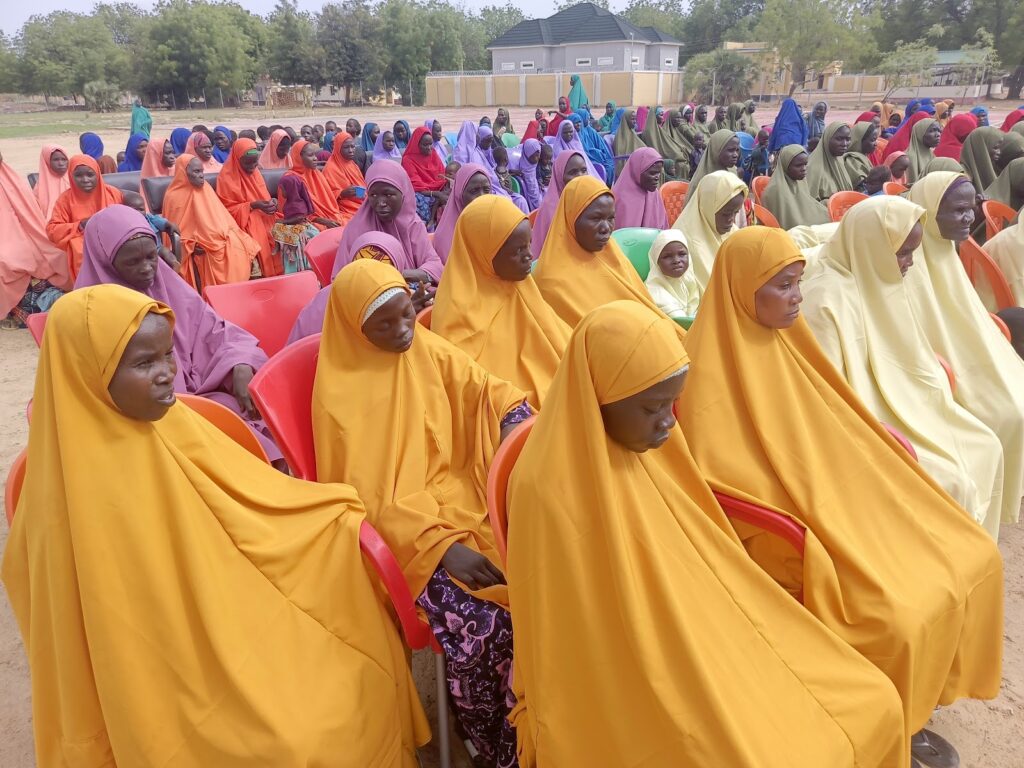 Group of women in colorful attire seated outdoors on plastic chairs, engaged in a community gathering.