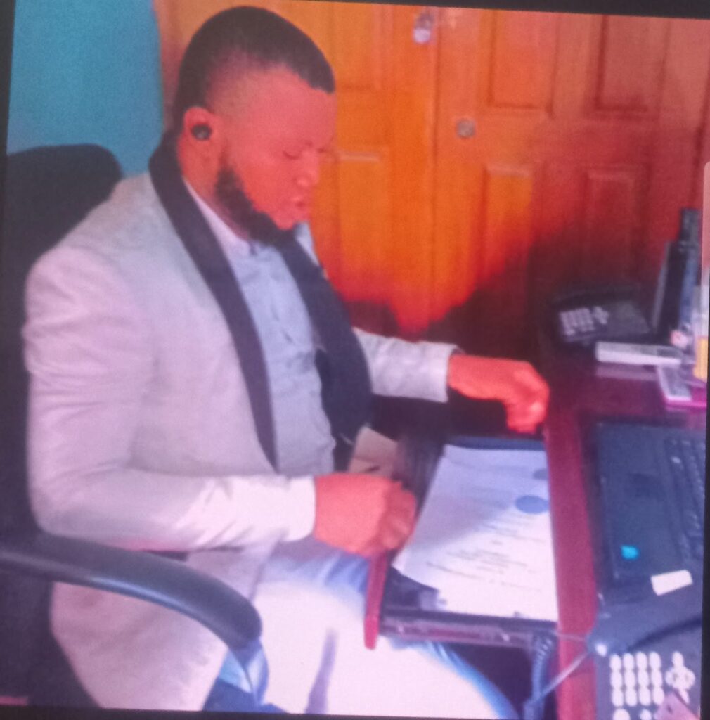 Man in a suit working at a desk with a laptop and documents, wearing a headset.