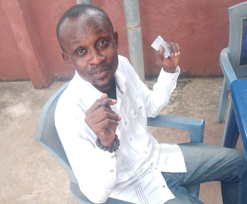 Man in white shirt sitting on a chair outdoors, gesturing with a small object in hand.