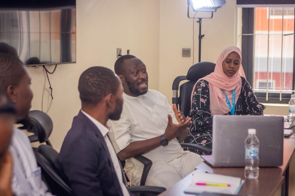 People in a meeting room with one man speaking and others listening, with a laptop and water bottles on the table.