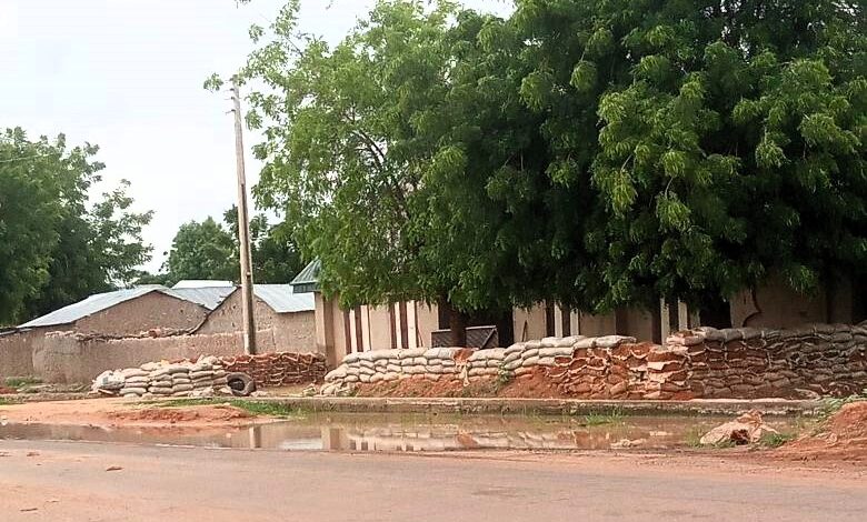 Rustic buildings with a sandbag barricade under leafy trees, near a puddle on a dirt road.