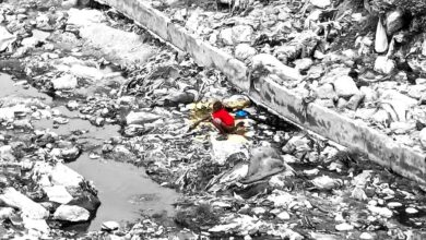 A selective color image of a person in red crouched amongst litter in a dry urban streambed.