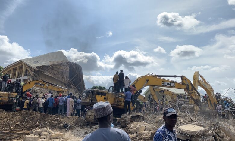 Collapsed building with onlookers and excavators amidst rescue efforts under a cloudy sky.