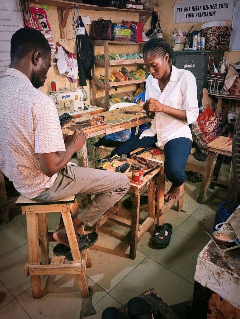 Two artisans crafting shoes in a busy workshop filled with tools and materials.