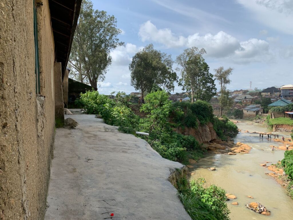 Rural landscape with a muddy river flowing past verdant foliage, near buildings under a cloudy sky.