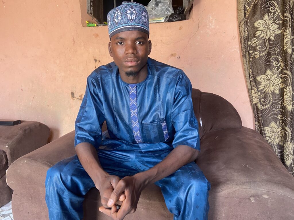 Man in blue traditional outfit sitting on a brown sofa with a patterned curtain in the background.