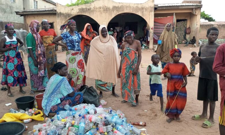 A group of people, including children, standing near a pile of plastic bottles in a village setting.