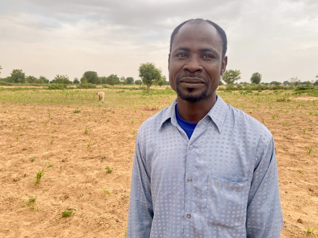 Man in blue shirt standing in a field with sparse vegetation under a cloudy sky.