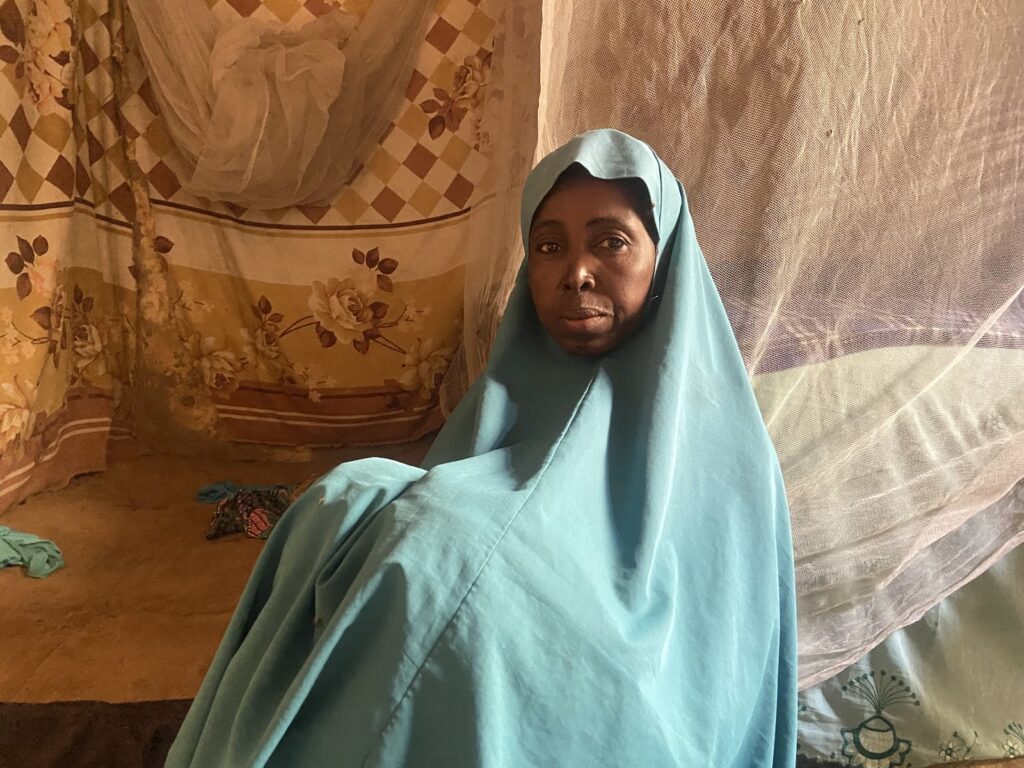 A woman draped in a blue shawl sits inside a room with patterned curtains and a mosquito net.