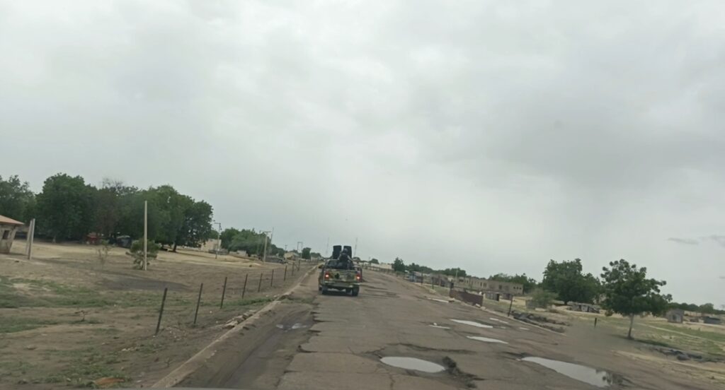 A vehicle driving on a potholed road with trees and buildings under an overcast sky.