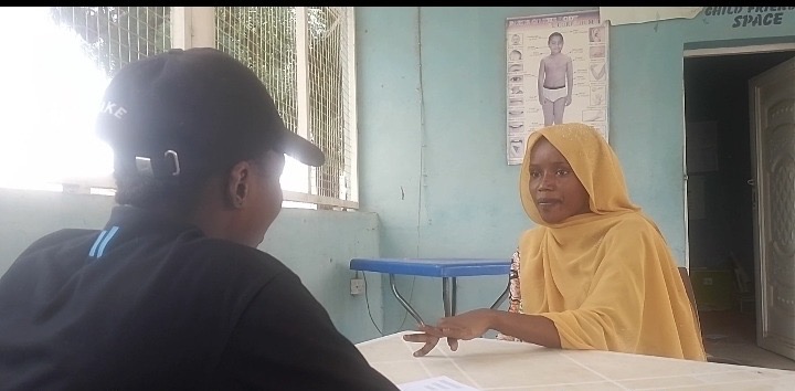 Two individuals engaged in a conversation across a table indoors, one in a baseball cap and the other in a yellow hijab.