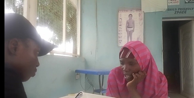 Two people sitting at a table in conversation, with posters on the wall in the background.