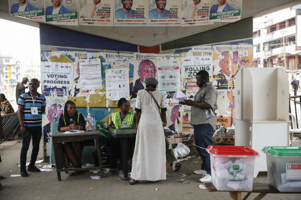 Voters at a polling station with officials and election materials, surrounded by political posters.