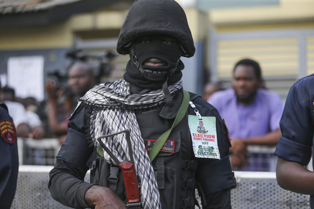 Masked security officer with an "Election Security Personnel" badge standing guard.