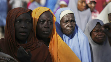 Group of women with contemplative expressions wearing colorful headscarves in a crowd.