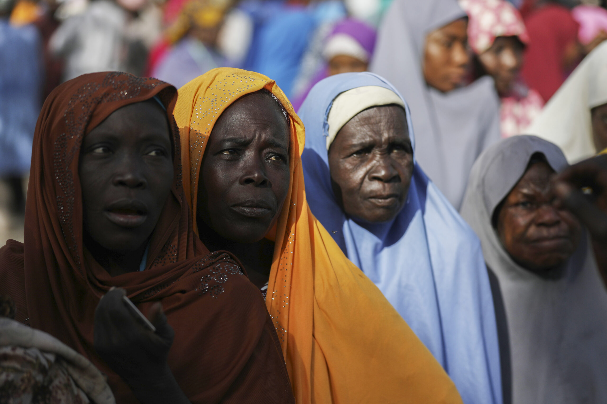 Group of women with contemplative expressions wearing colorful headscarves in a crowd.