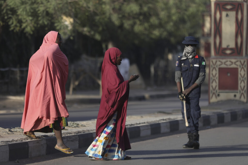 Two women in traditional attire walking past a masked security officer on a street.