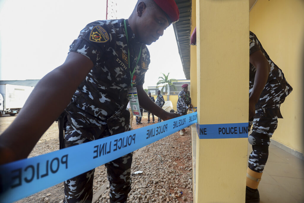 An officer in camo erects a police line tape at a cordoned area while colleagues observe in the background.