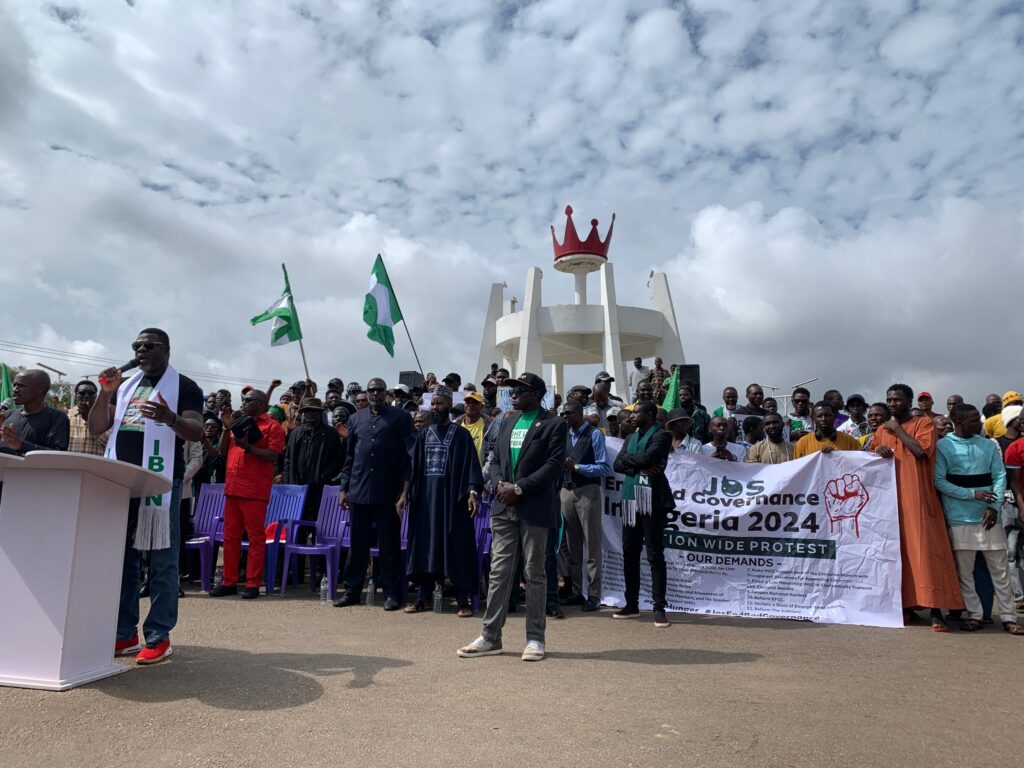 Group of people at a rally with banners, one reading "Jobs and Governance Nigeria 2024", under a cloudy sky.