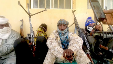 Group of armed individuals in camouflage, with covered faces, sitting against a wall.