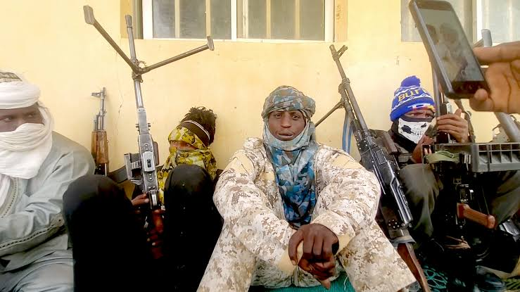 Group of armed individuals in camouflage, with covered faces, sitting against a wall.