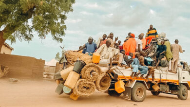 Overloaded truck with people and goods on a dusty road, under a blue sky with a tree in the background.