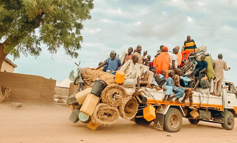 Overloaded truck with people and goods on a dusty road, under a blue sky with a tree in the background.