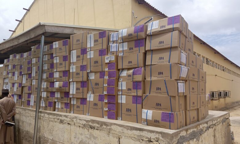 Stacks of cardboard boxes labeled "IDA" outside a building, with two people standing nearby.