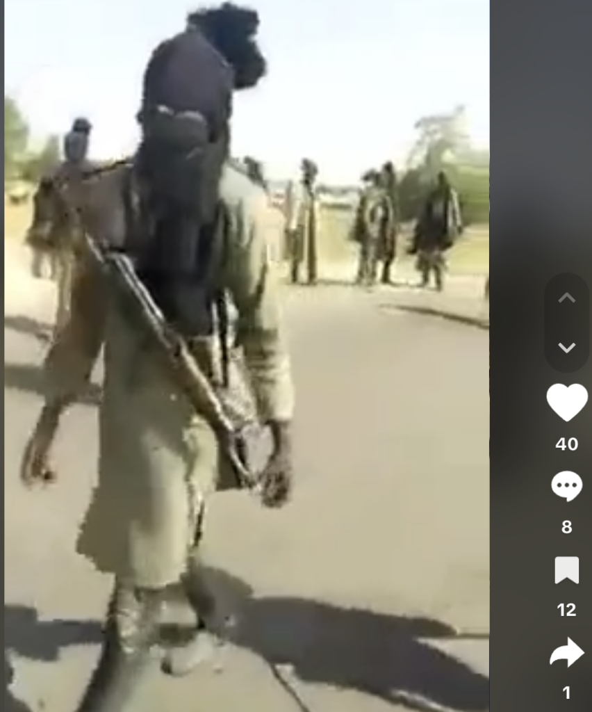 Masked person with a firearm walking, others in the background on a sunny street.