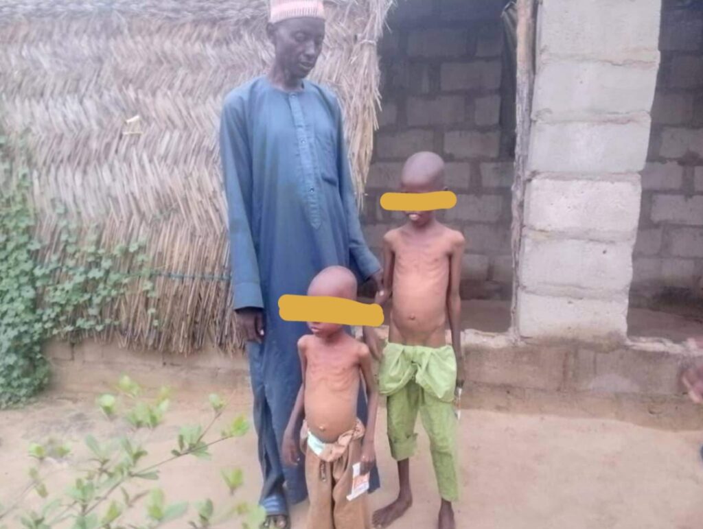 Man in traditional attire standing with two children outside a house with a thatched roof.