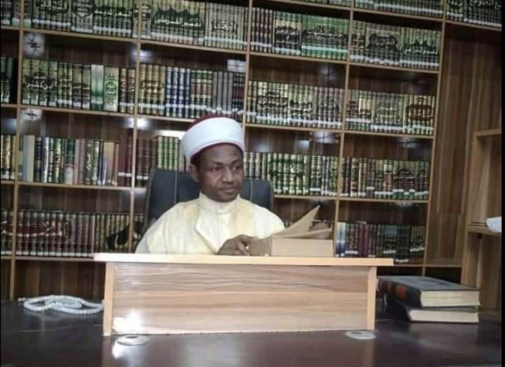 Man in a traditional cap reading a book at a desk with a large bookshelf filled with books behind him.