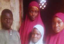 Four people posing, three in red hijabs, and one boy in a grey shirt, standing in front of an old wall with a lattice window.