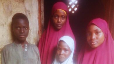 Four people posing, three in red hijabs, and one boy in a grey shirt, standing in front of an old wall with a lattice window.