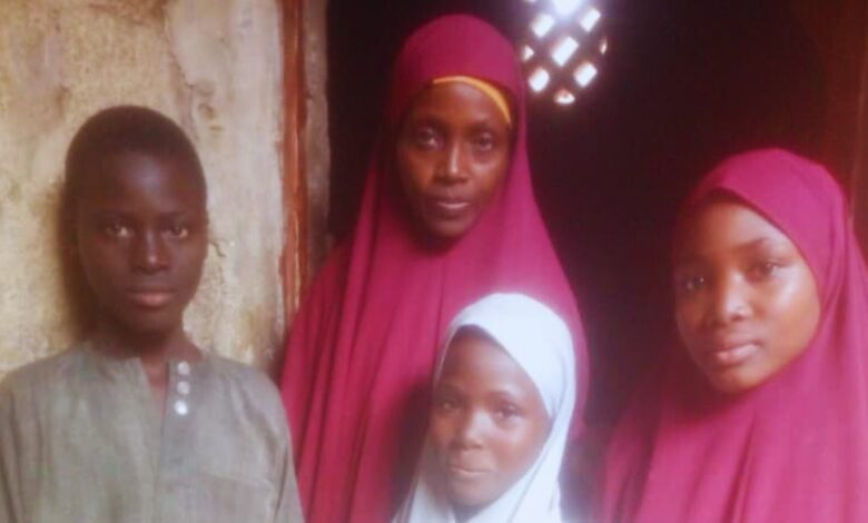 Four people posing, three in red hijabs, and one boy in a grey shirt, standing in front of an old wall with a lattice window.