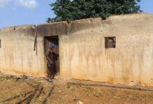 Person stands at the entrance of a weathered building with a damaged roof and a broken window.