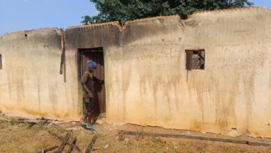 Person stands at the entrance of a weathered building with a damaged roof and a broken window.