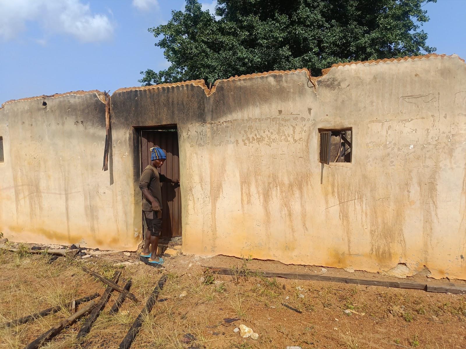 Person stands at the entrance of a weathered building with a damaged roof and a broken window.